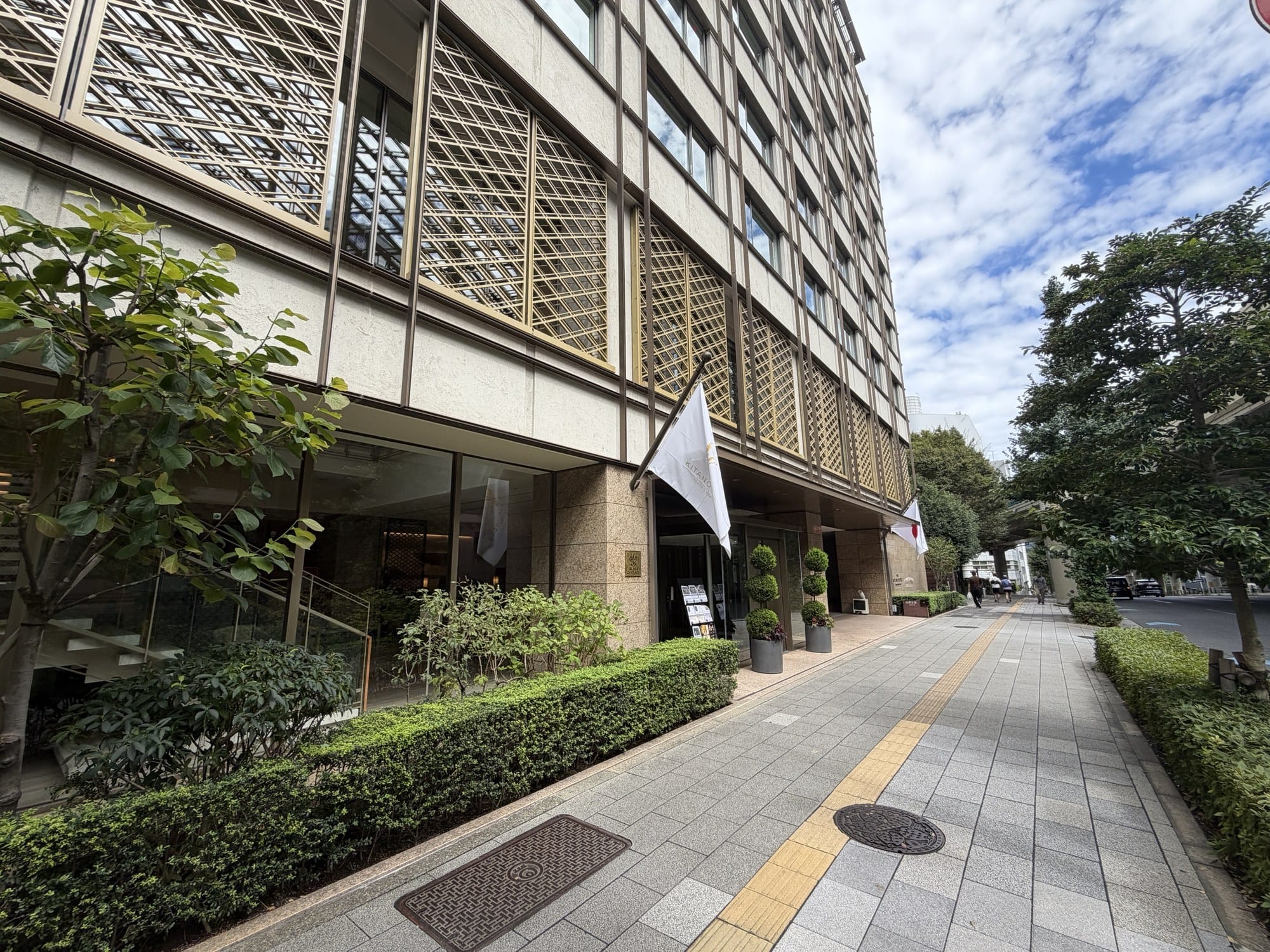 The Kitano Hotel Tokyo seen from the sidewalk, with the gold lattice screen covering the upper floors and trimmed street hedges out front.