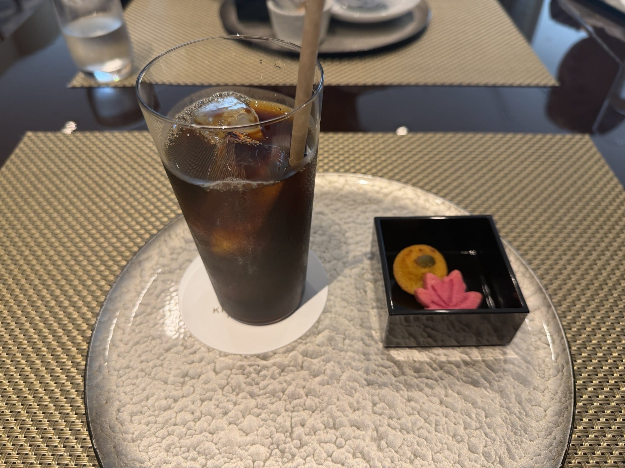 Iced coffee in a tall glass with a paper straw and large ice cubes, next to a black lacquer box holding two gluten-free rice-flour sweets