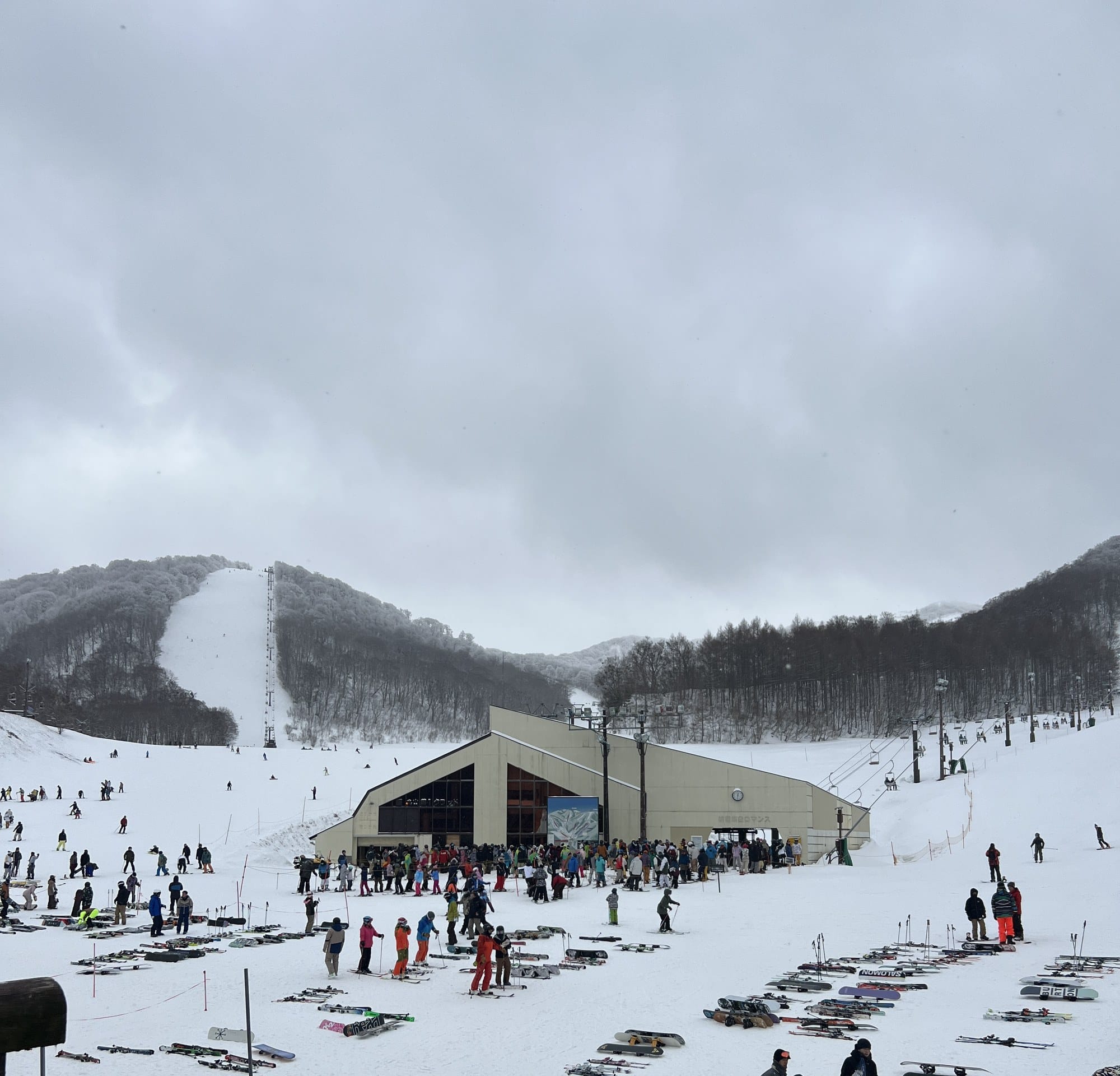 Skiers and snowboarders gathered outside the north base lodge at Nekoma Mountain, with rime-covered beech forest and pisted runs behind