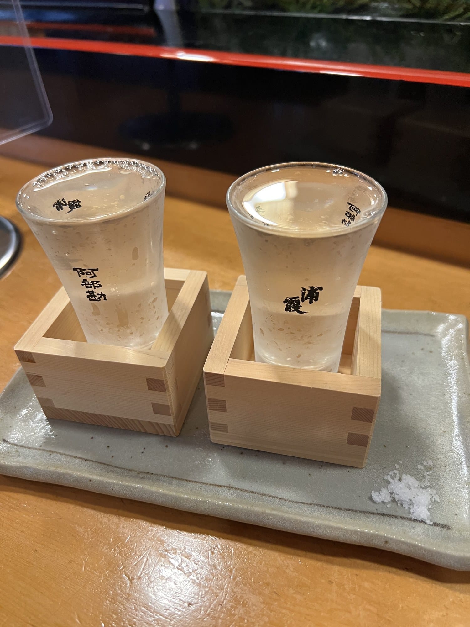 Two chilled sake glasses, one labeled Urakasumi and one labeled Abekan, served on wooden trays with a small mound of salt