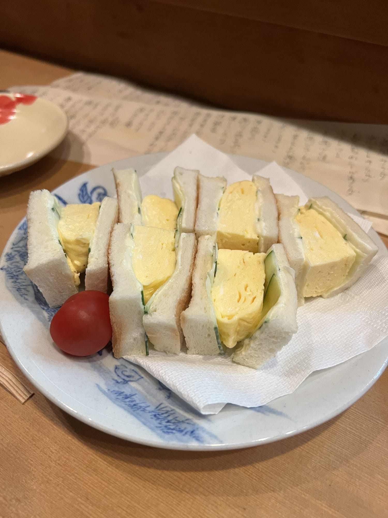 Egg sandwiches of thick rolled omelette and cucumber on white bread, on a blue-and-white plate with one cherry tomato, at a wooden counter