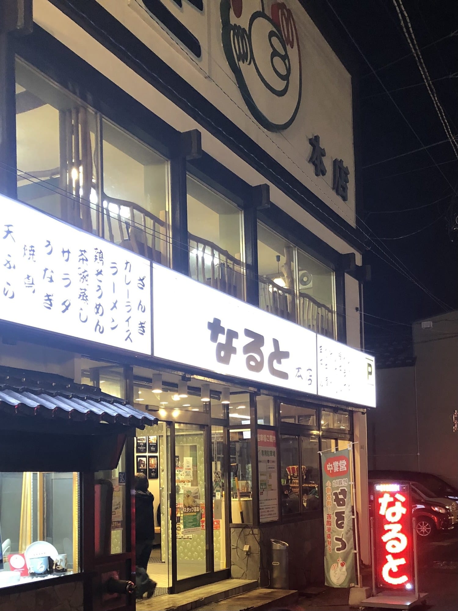 The lit exterior of Naruto's honten restaurant at night in Otaru, with its menu board and a parking sign