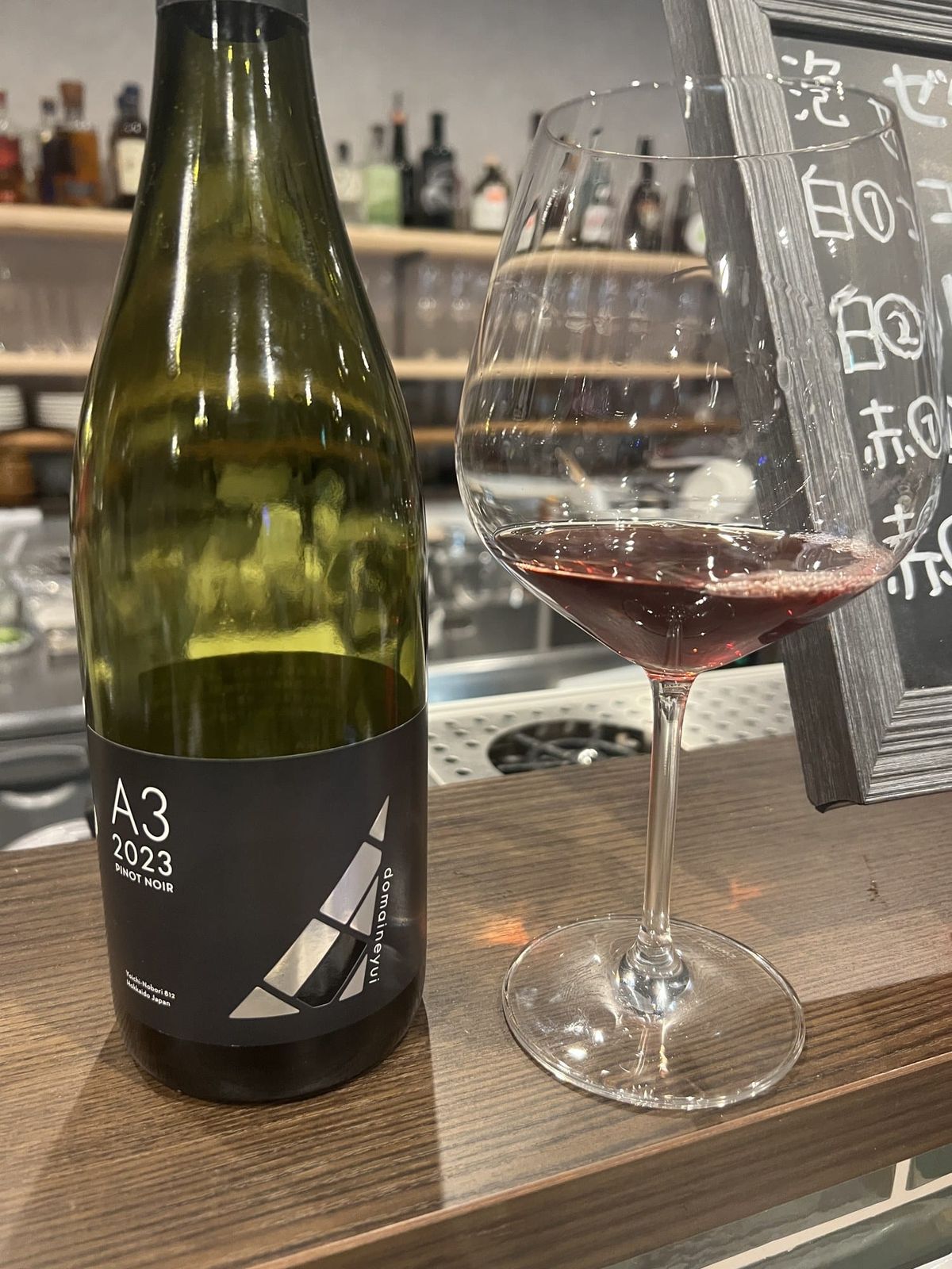 A glass of red wine and a bottle of Domaine Yui A3 2023 on a Yoichi wine bar counter