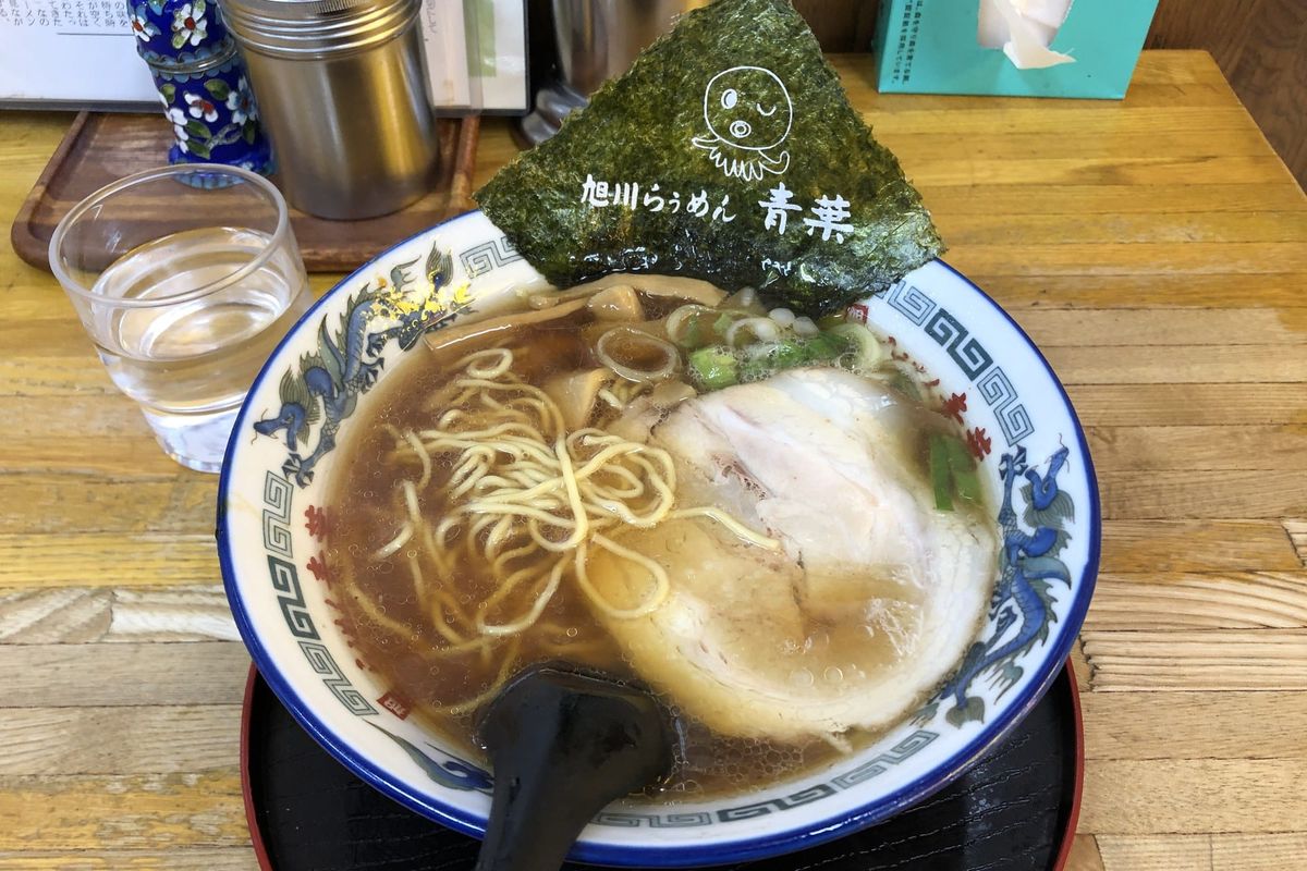A bowl of Asahikawa soy-sauce ramen with two slices of chashu, bamboo shoots and a sheet of nori, on a wooden counter
