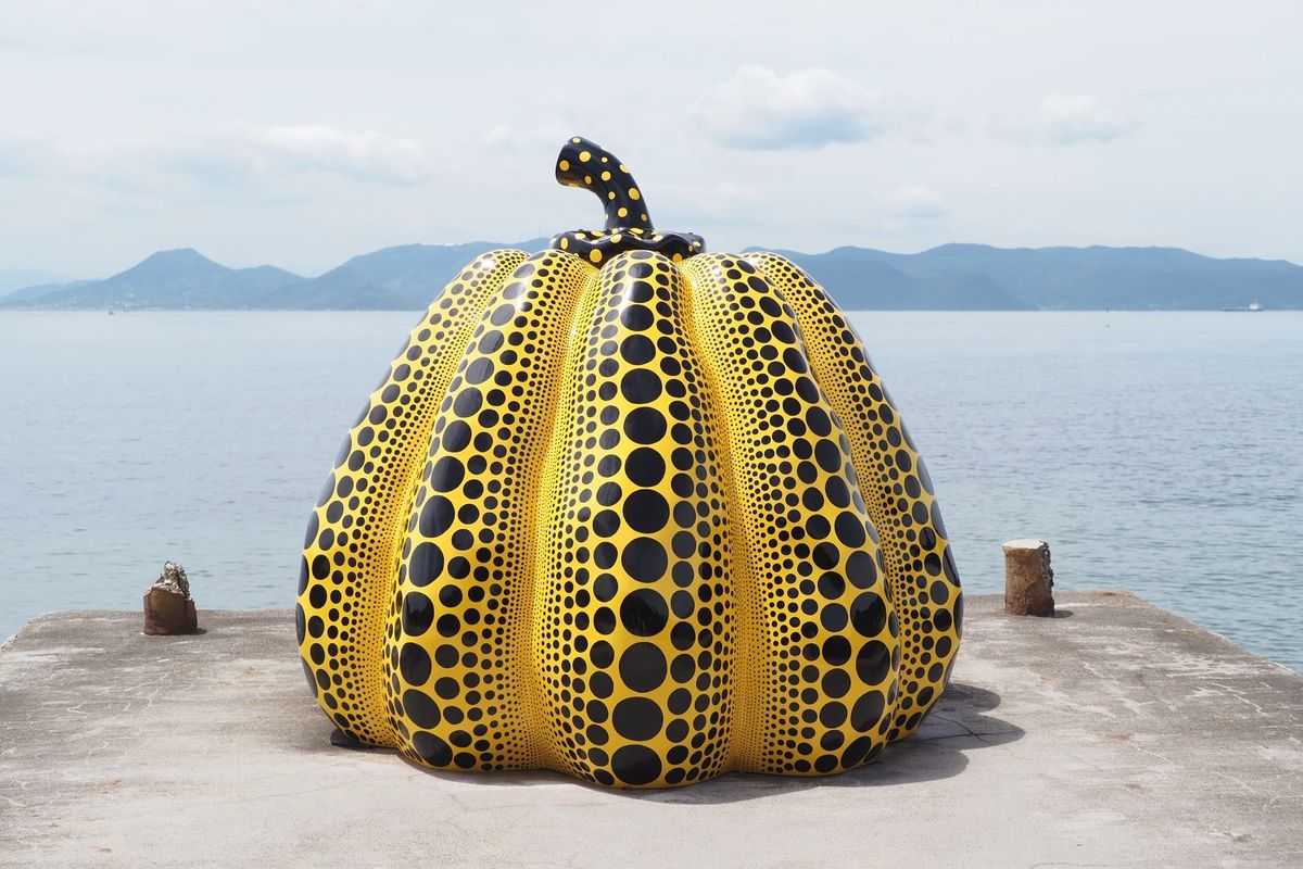 A large yellow spotted pumpkin sculpture on a concrete pier, with the Seto Inland Sea and distant islands behind it