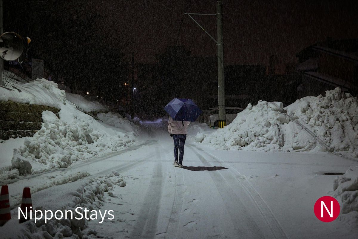 A snow-banked street in Fukui city at night, a figure walking under an umbrella