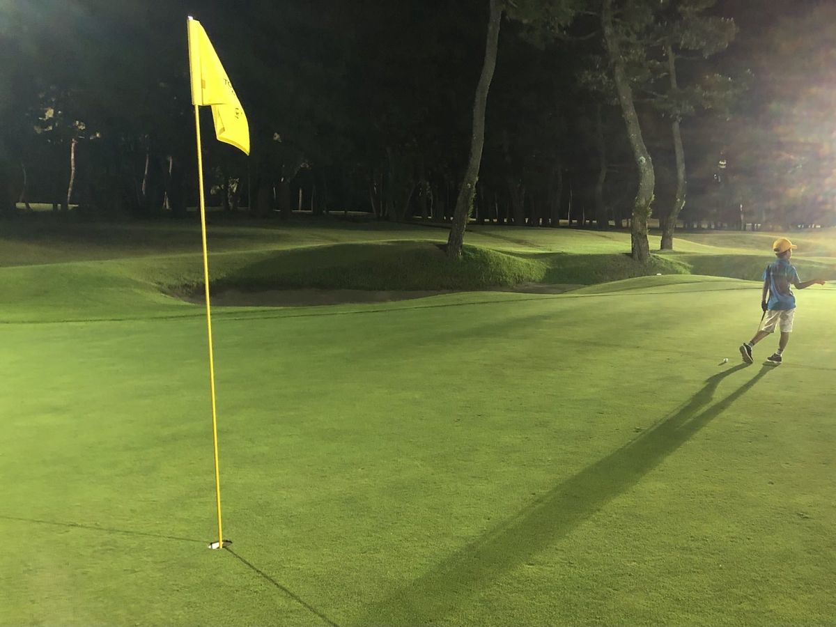 A child walking up a floodlit green at night on the Tom Watson Golf Course, long shadow cast ahead