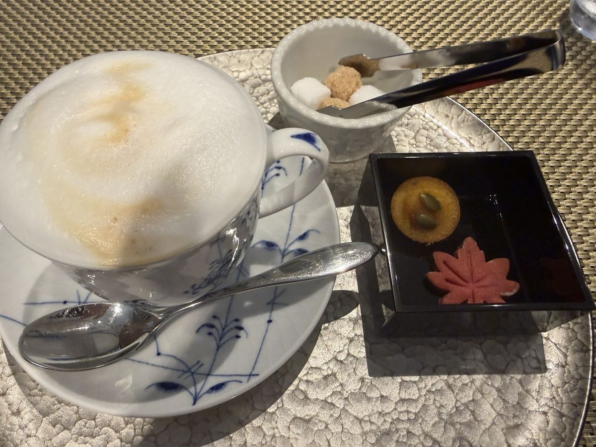 Café au lait with thick foam in a blue-and-white cup, beside a black lacquer box of two gluten-free rice-flour sweets