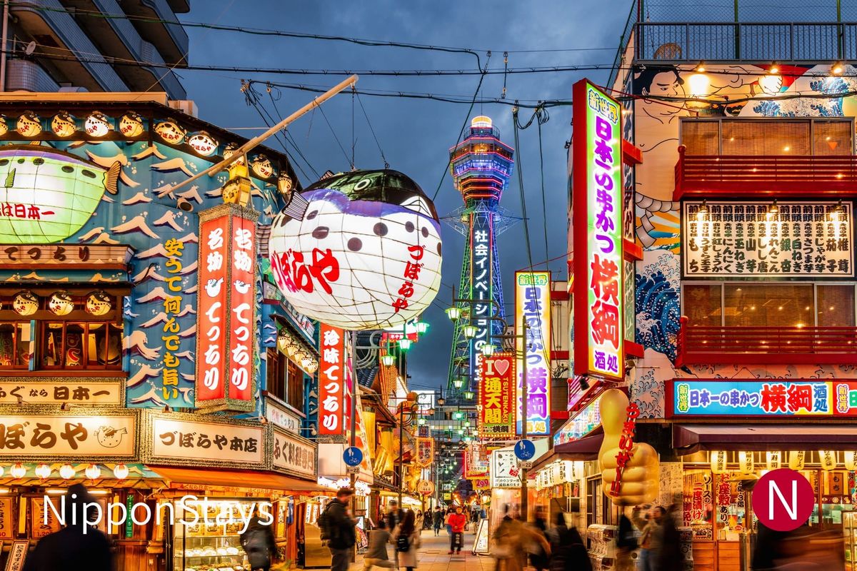 Shinsekai street at dusk under lanterns and neon signs, with Tsutenkaku Tower lit behind