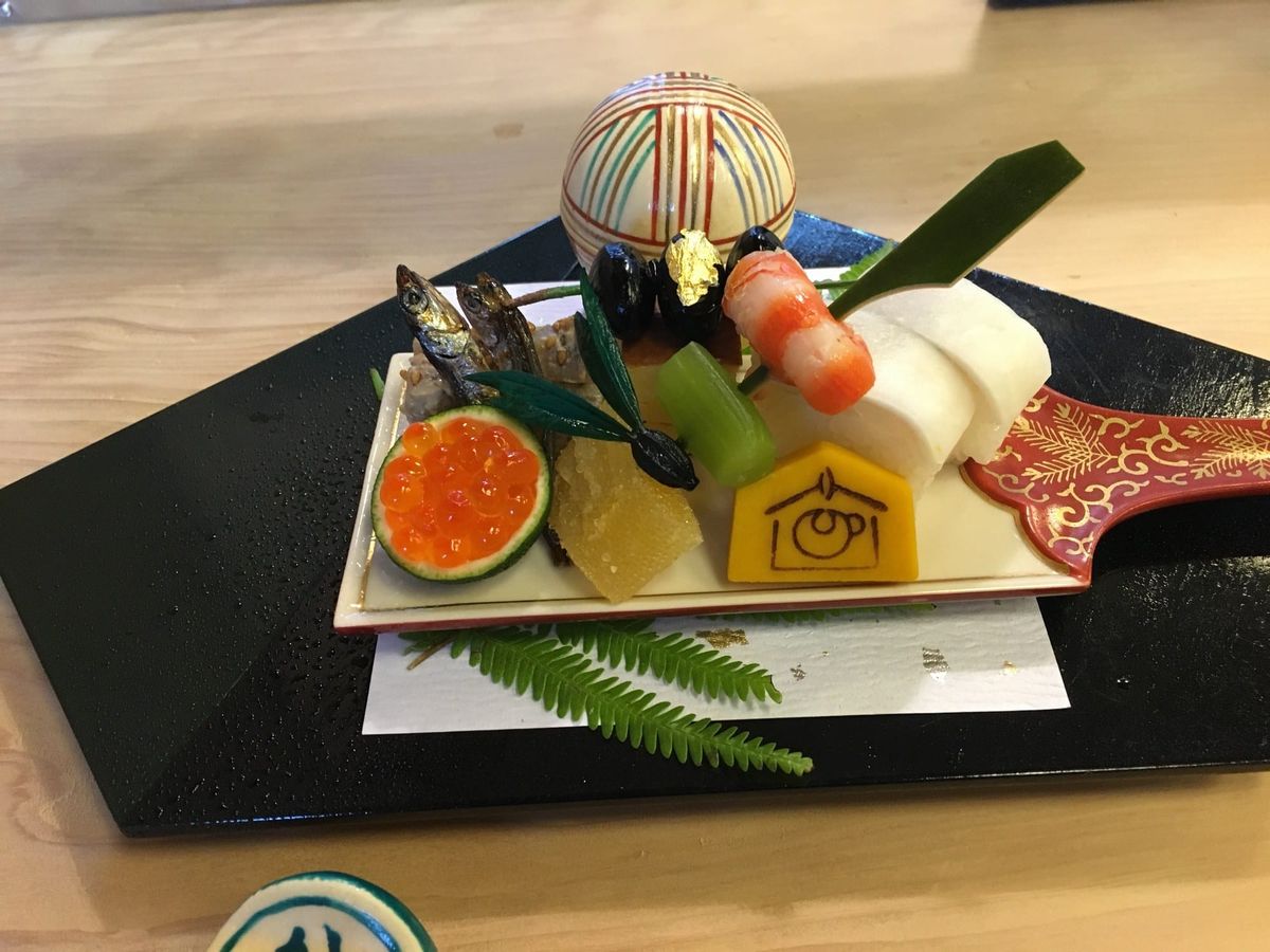 A New Year sakizuke course on a black lacquer board: black beans with gold leaf, small fish, salmon roe in a hollowed citrus, a prawn roll and a temari sweet, on fern
