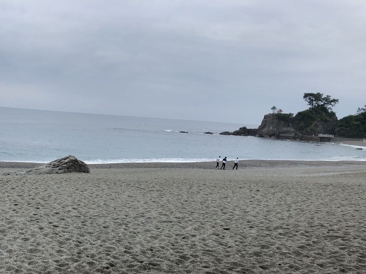 Katsurahama beach in Kochi under a grey sky, with a pine-topped headland and a small red shrine pavilion at the far end of the bay