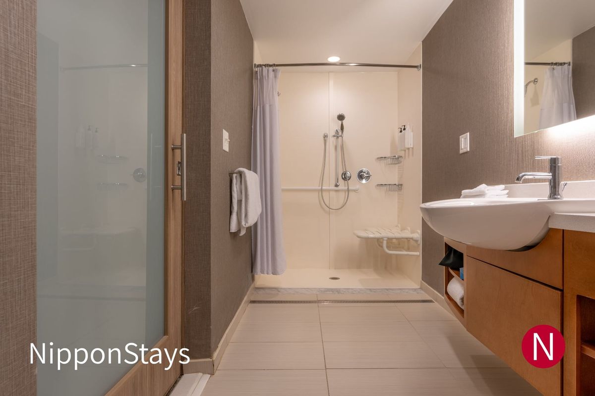 A roll-in shower with a fold-down seat and grab bars in an accessible hotel bathroom