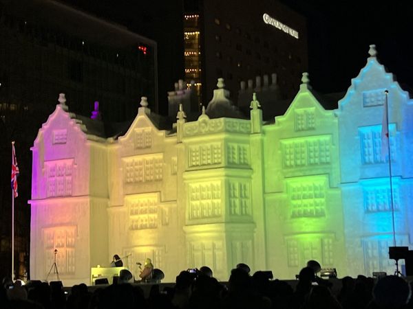 A large illuminated snow sculpture of a European building facade lit in rainbow colors during the Sapporo Snow Festival
