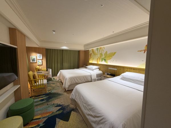 Hotel room with two beds, a low table and armchairs, and a leaf-patterned mural above the headboard
