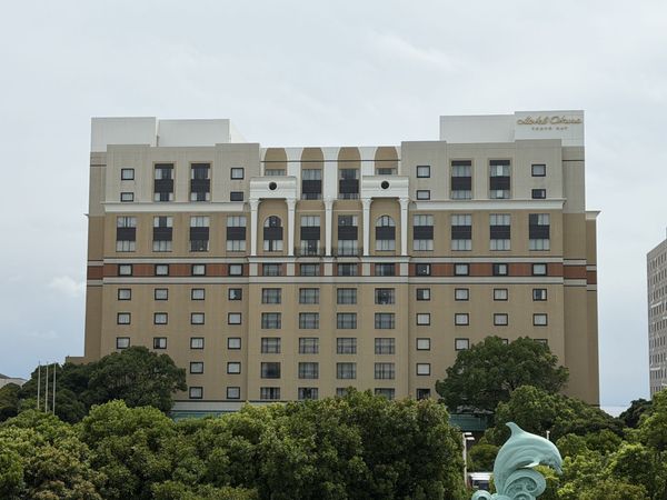 The beige facade and rooftop sign of Hotel Okura Tokyo Bay, seen above the trees from the Bayside hotel area
