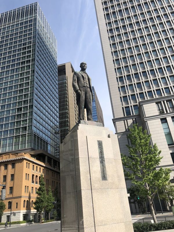 Bronze statue of Inoue Masaru in a Western suit on a tall granite pedestal, with the Marunouchi office towers of Tokyo Statio
