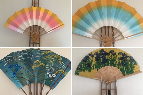 Four Kyoto folding fans opened on wooden stands, in orange-pink, orange-blue, painted blue autumn grasses and yellow with iri