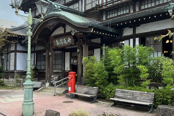 The curved gable entrance of Takegawara Onsen in Beppu, a wooden municipal bathhouse, with a red postbox, two benches and a s