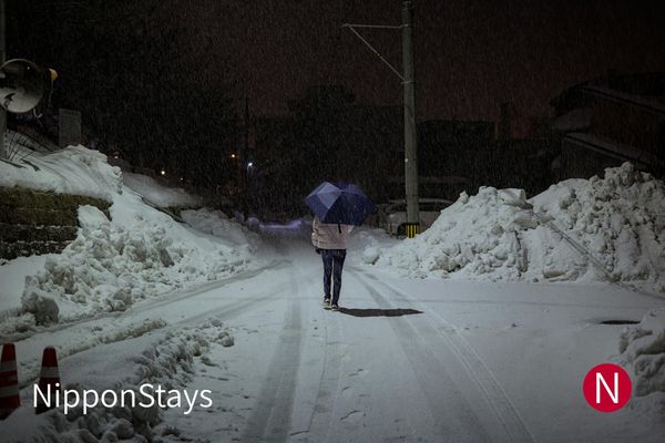 A snow-banked street in Fukui city at night, a figure walking under an umbrella