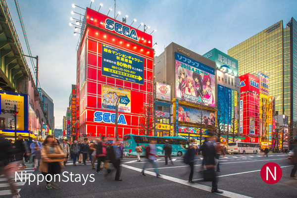 Pedestrians crossing a broad street beneath Akihabara's electronics signs.