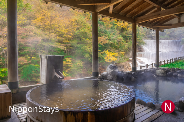 An empty open-air Japanese onsen bath beside a wooded waterfall.