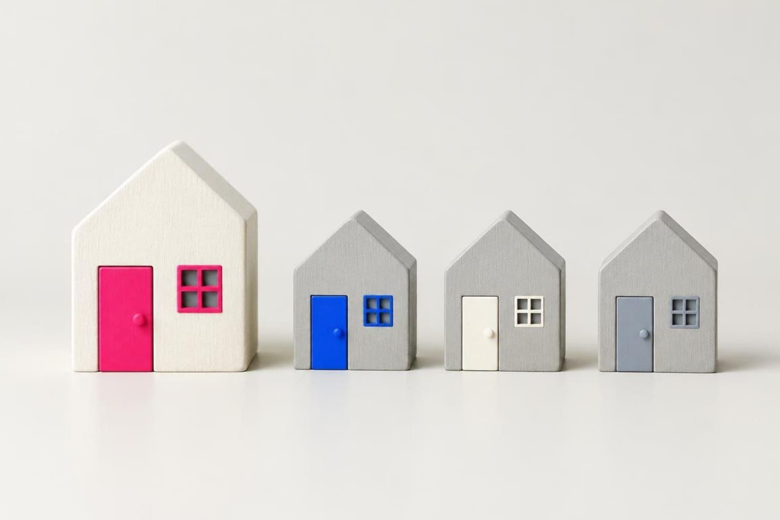 Four small wooden houses in a row: a larger white one with a pink door, then three grey ones with blue, white and grey doors.