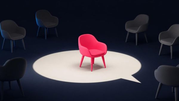 One pink chair stands in a spotlight shaped like a speech bubble, with empty grey chairs in the dark around it.