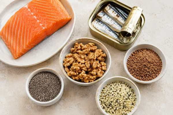 Omega-3 food sources including salmon, sardines, walnuts, chia seeds, flaxseed, and hemp seeds arranged on a light background.