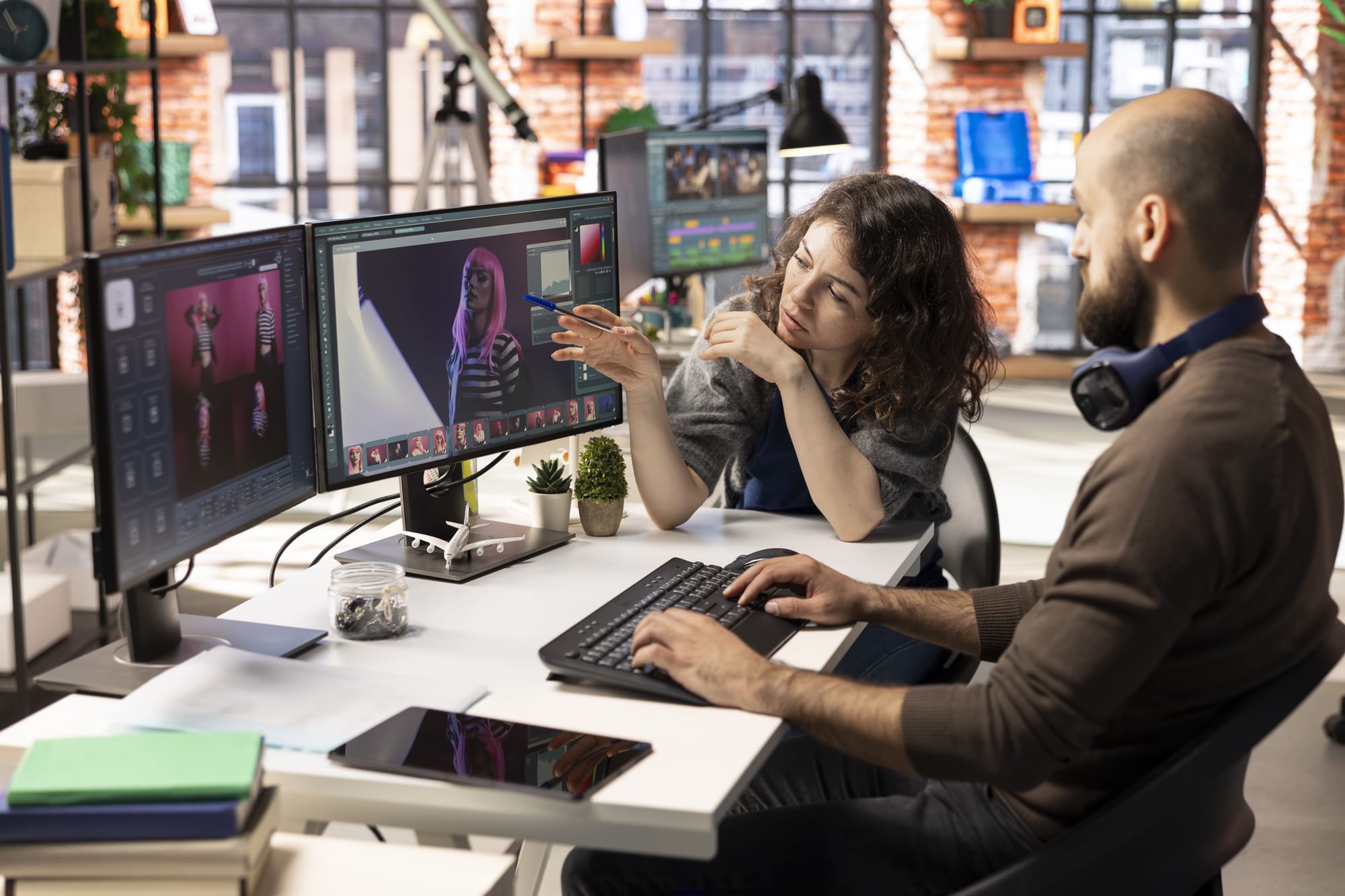 Two colleagues at a desk in a studio, discussing an AI-generated portrait of a pink-haired model displayed on a large monitor.