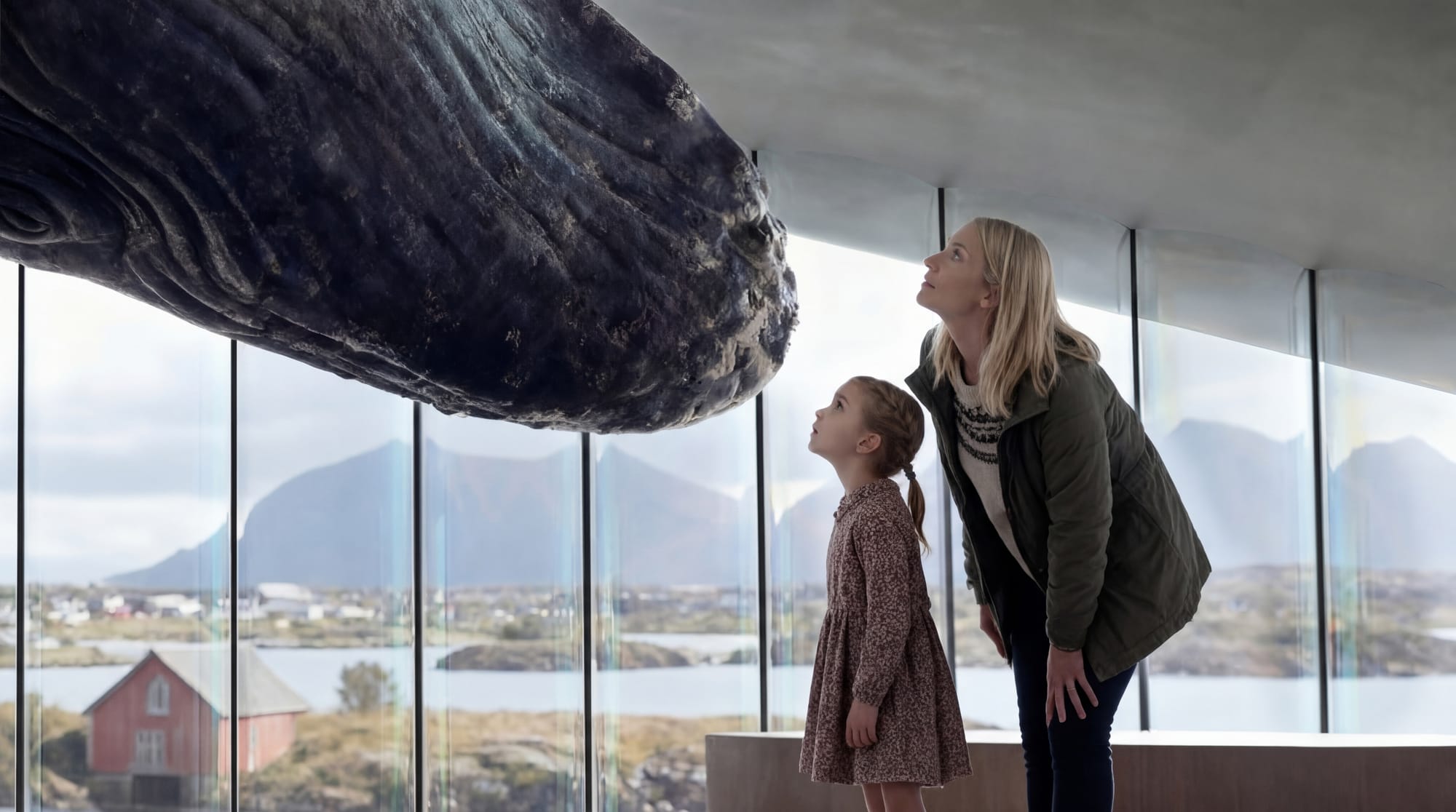 A mother and daughter look up at the underside of a large sculpted whale tail suspended from the ceiling, with a coastal Norwegian landscape visible through floor-to-ceiling windows behind them.