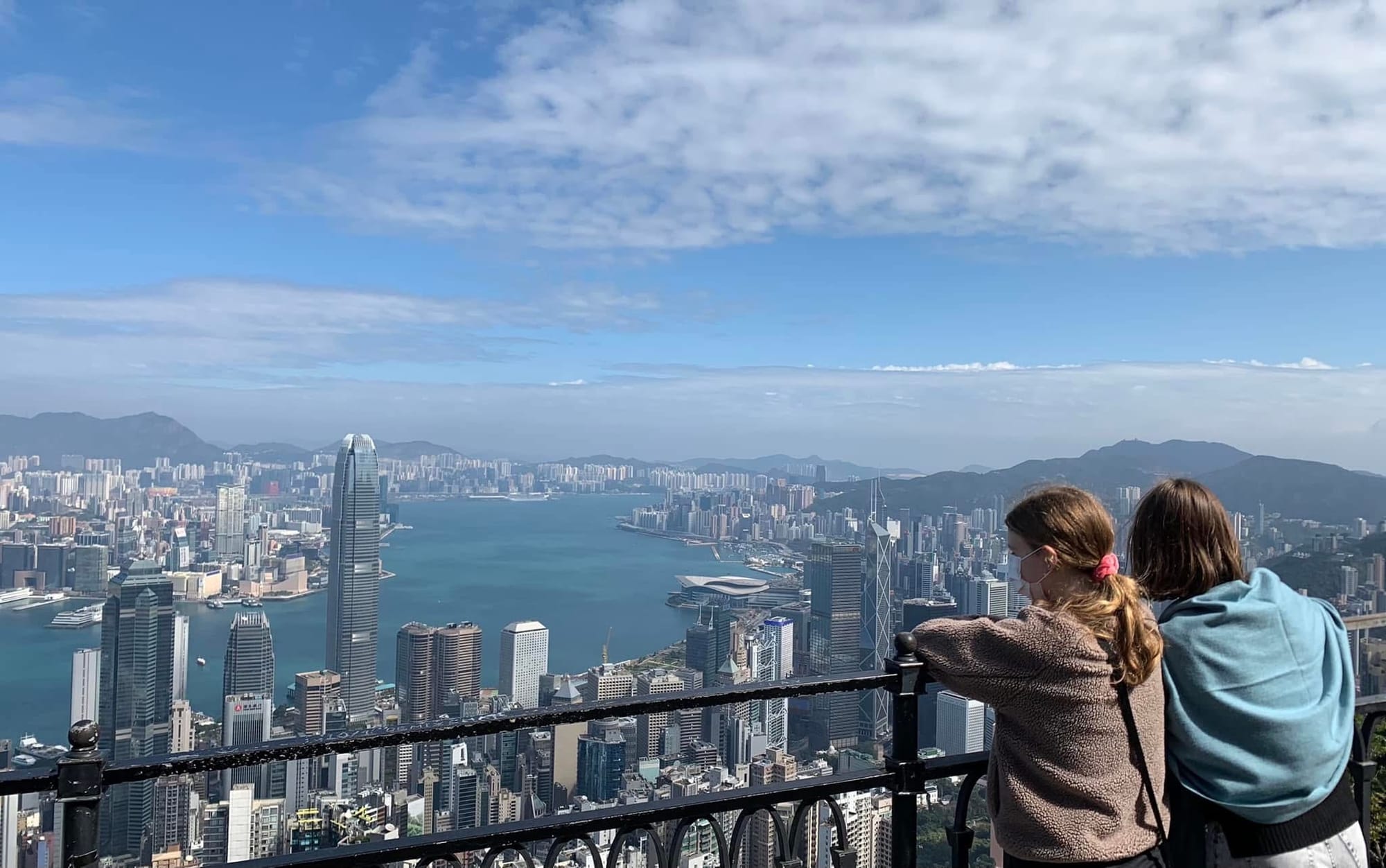 Two women viewed from behind standing at a lookout railing on Victoria Peak, gazing across the Victoria Harbour skyline and mountains of Hong Kong on a clear day.