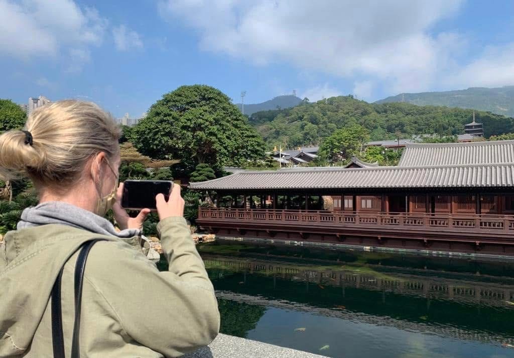 A woman viewed from behind wearing a mask and jacket, using her smartphone to take a photo of a traditional Chinese courtyard building and koi pond at Nan Lian Garden.
