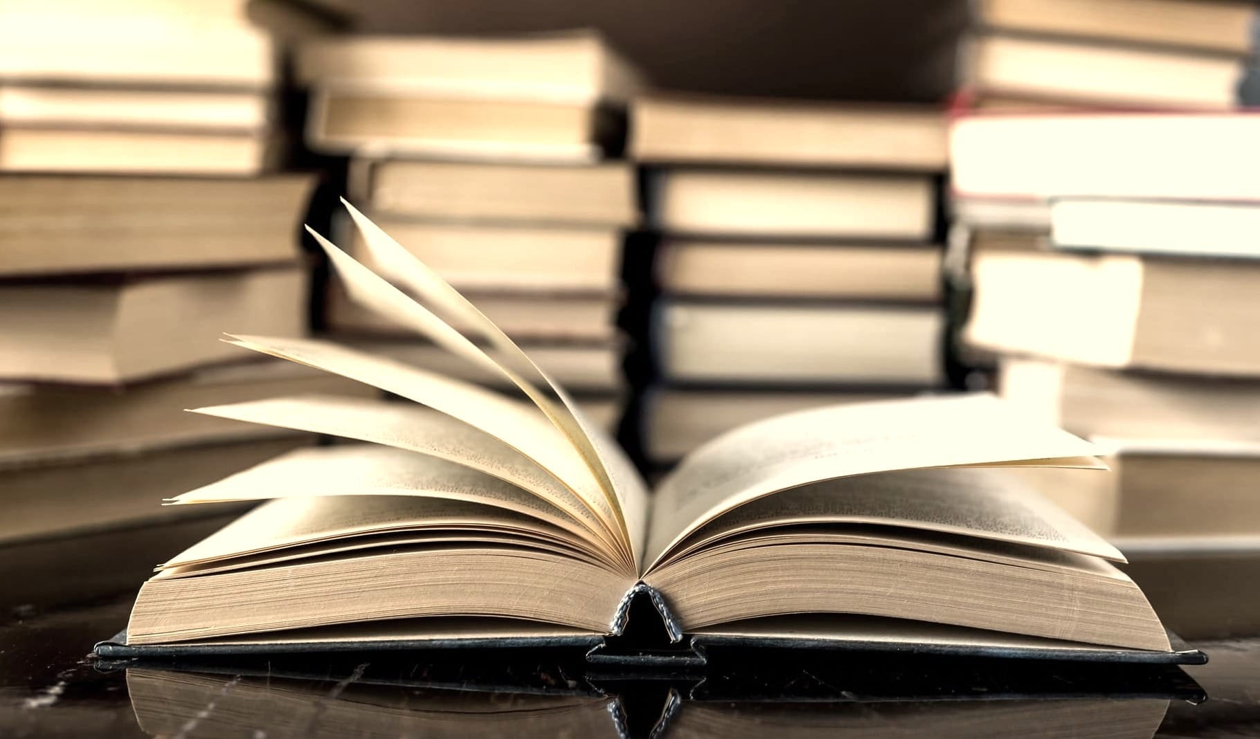 An open hardcover book with fanned pages resting on a dark reflective surface, surrounded by tall stacks of closed books.