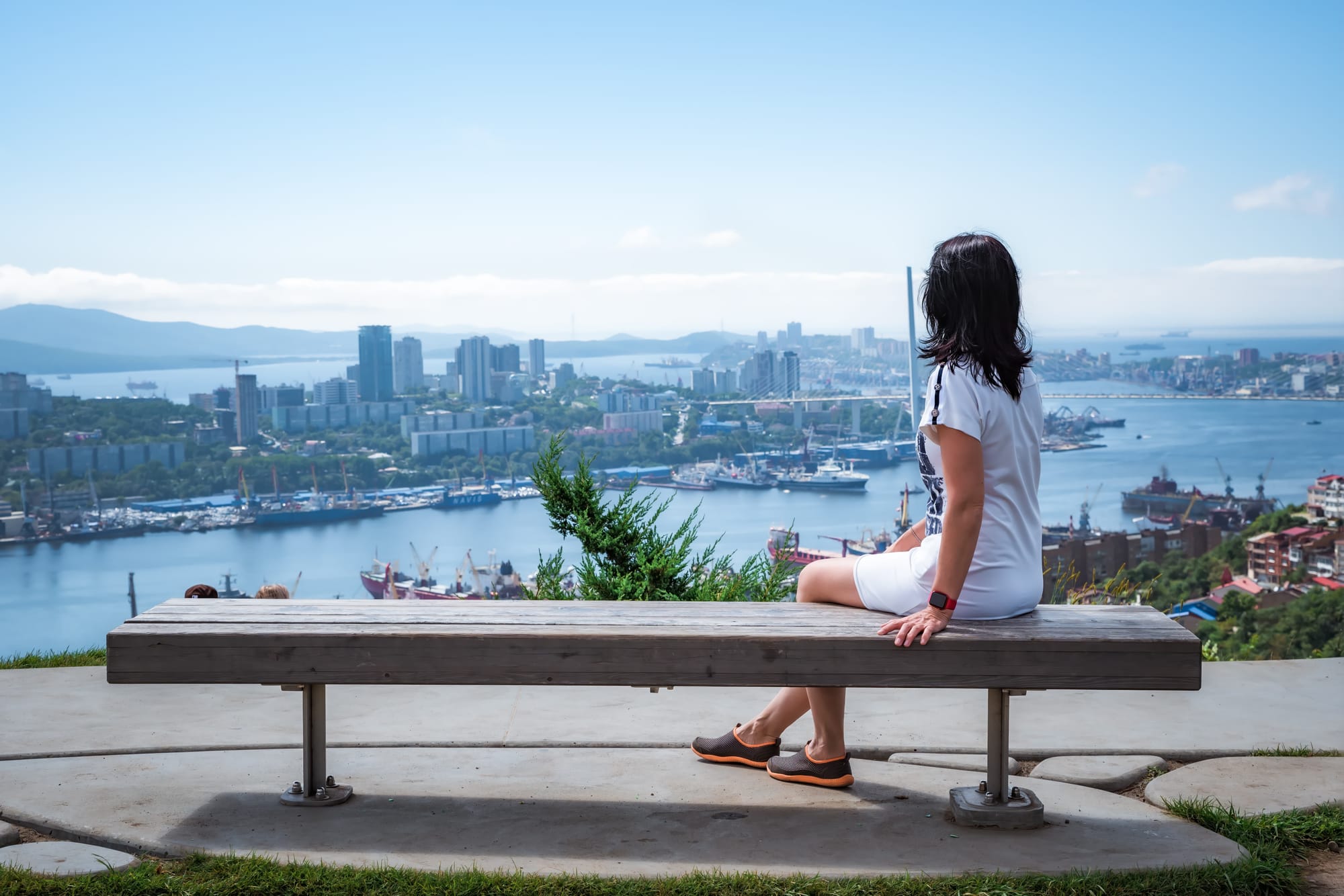Woman sitting alone on a bench overlooking a harbour city, facing away from the camera toward the view