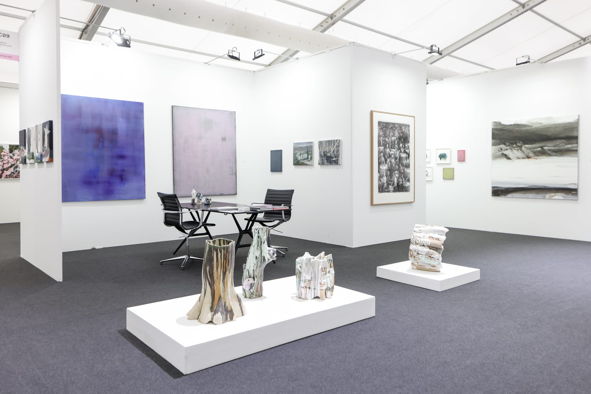 A white-walled gallery booth at an art fair displaying abstract paintings, framed prints, and ceramic sculptures on plinths.