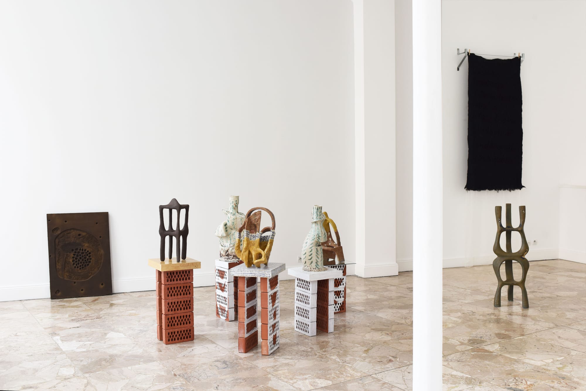 Ceramic sculptures on brick pedestals in a gallery, with a black woven textile piece hanging on the wall behind.