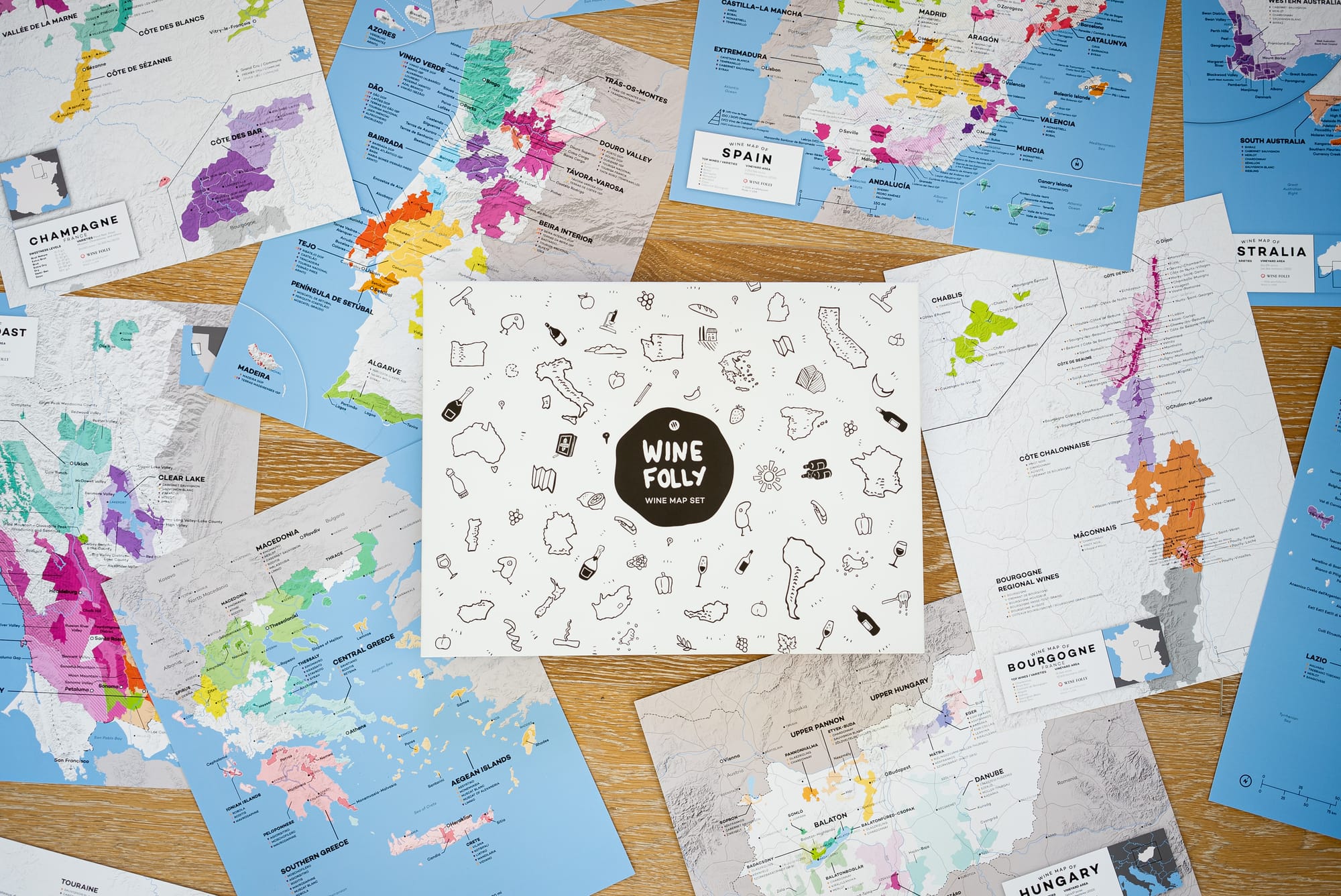Flat-lay view of the central "Wine Folly Wine Map Set" box surrounded by detailed, colorful maps of world wine regions on a light wooden table.