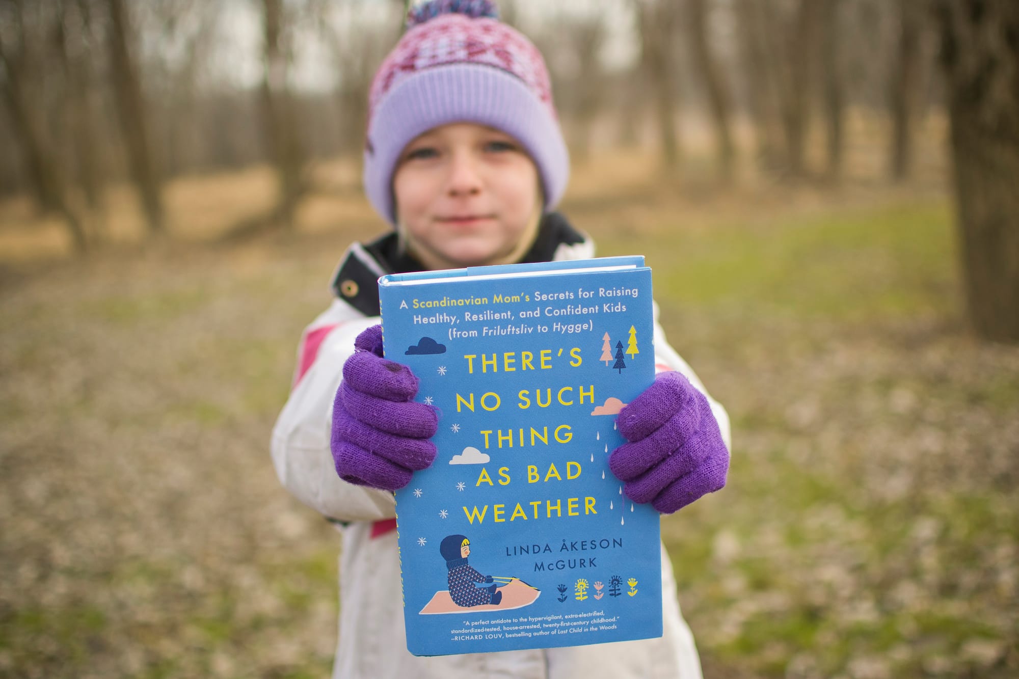 A young girl in a purple knit hat and gloves holds out a blue hardcover book titled There's No Such Thing as Bad Weather in a bare, wooded park.