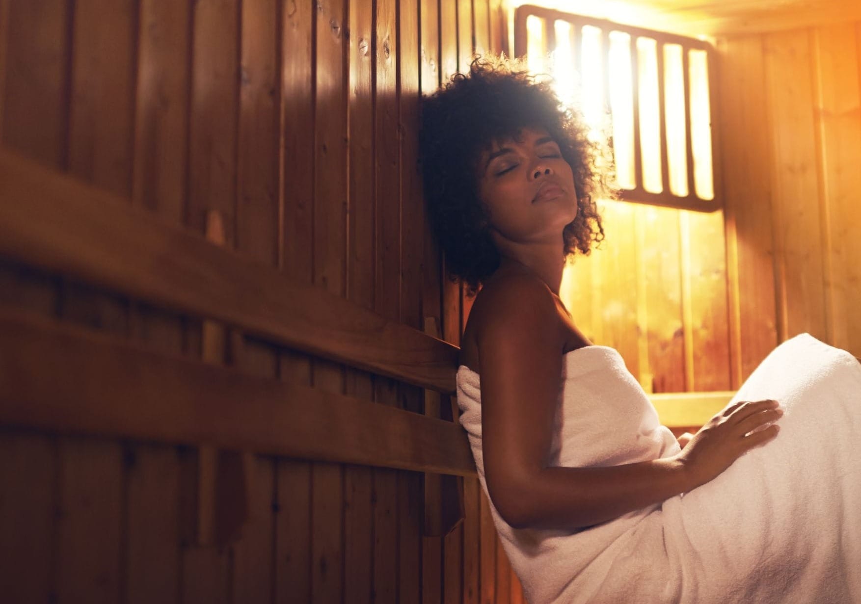 A woman wrapped in a white towel sits with her eyes closed, resting against the wooden wall of a warmly lit sauna.