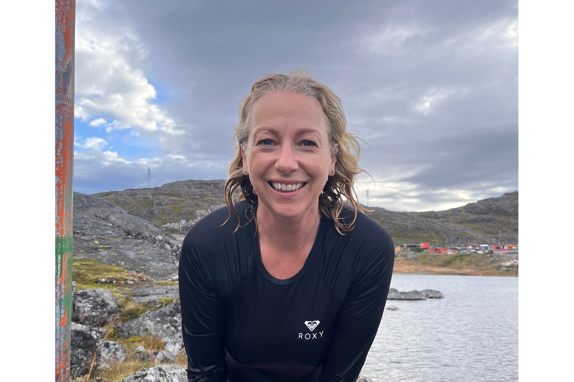 Laura Hall, wet-haired and smiling, in a long-sleeved black swim top beside a lake, with rocky hills and colourful houses behind her.