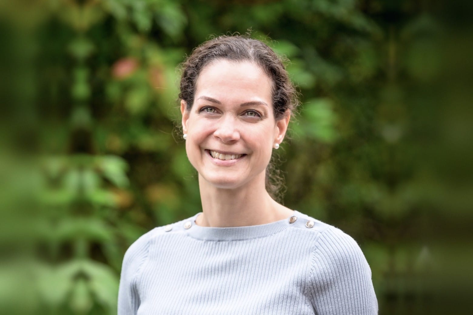 Dr Wendy Knoops, a woman with dark hair tied back, smiling and wearing a light blue jumper in front of green foliage