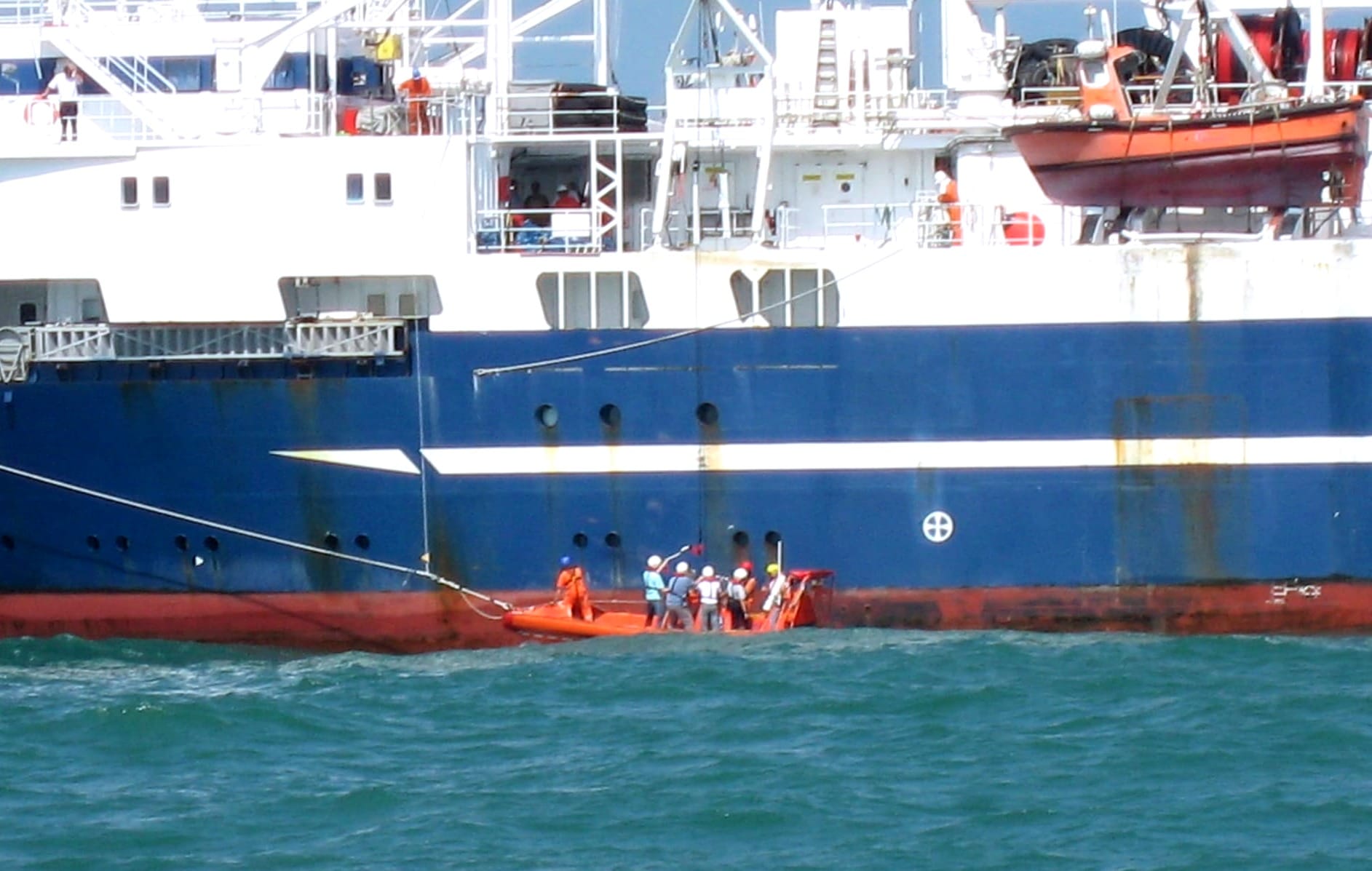 A small orange rescue boat with crew in hard hats and life jackets alongside a large blue and white vessel, on choppy green-blue sea off the coast of China.