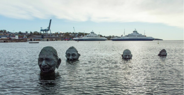Four large bronze heads by Bruce Naigles rising out of the sea in Moss harbour, with ferries in the background.