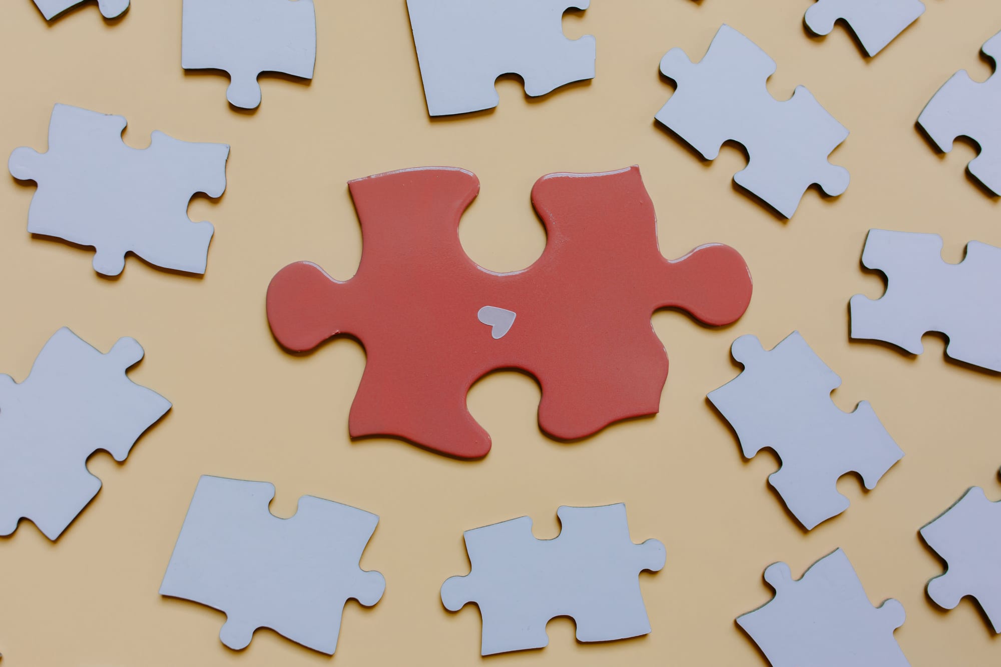 A single red puzzle piece with a small white heart, surrounded by scattered white puzzle pieces on a warm yellow background.