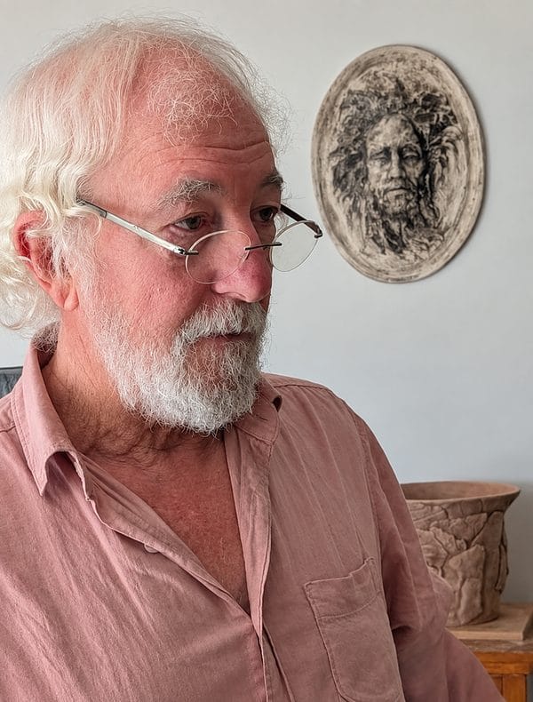 Portrait of sculptor Bruce Naigles with white hair, beard and glasses, wearing a pink shirt, with a carved relief of a face on the wall behind him.