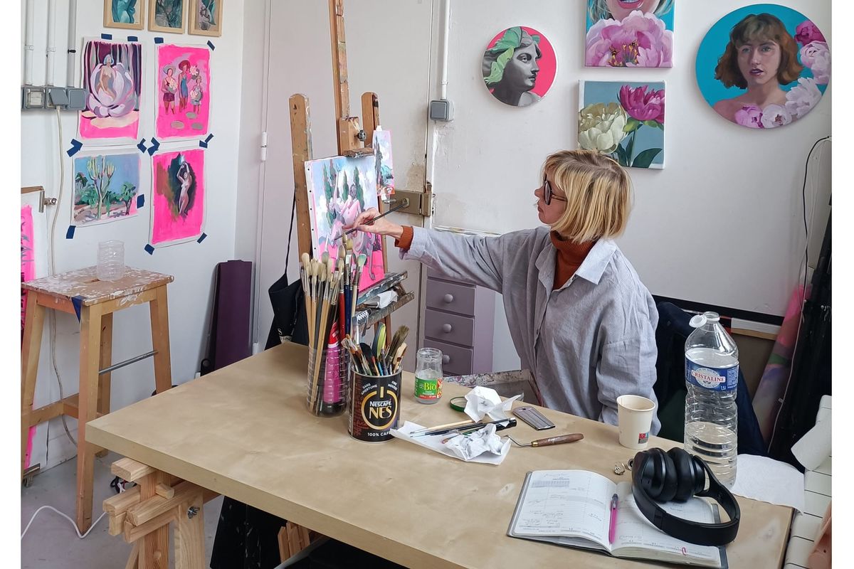 Liz Adams, a blonde woman in glasses and a striped shirt, paints at a wooden easel in a white studio with colourful canvases on the walls.