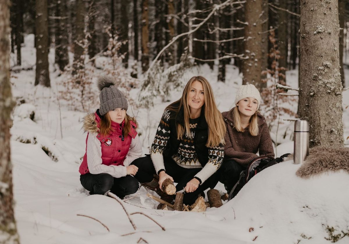 A woman in a Nordic-patterned knit sweater whittles a log in a snowy spruce forest, crouching between her two daughters in winter hats, with a thermos and backpack beside them.