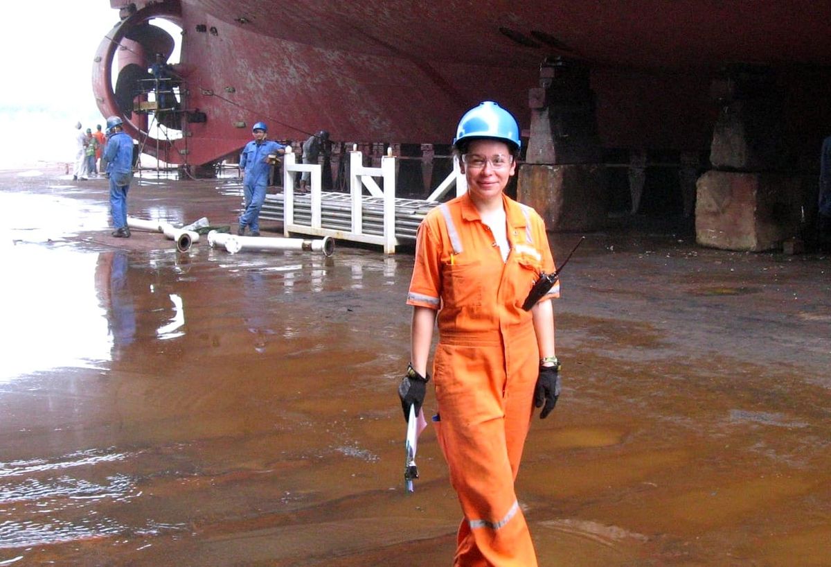 Aurelia Paraschiv in orange coveralls and a blue hard hat, standing in a dry dock beneath the hull of a ship.