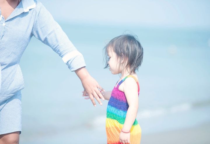 A child's hand reaching toward her mother's outstretched hand, both blurred against a soft blue sea and sky.