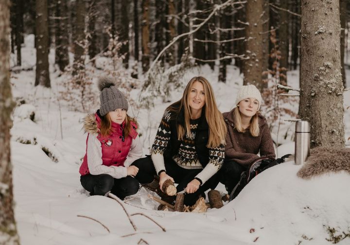 A woman in a Nordic-patterned knit sweater whittles a log in a snowy spruce forest, crouching between her two daughters in winter hats, with a thermos and backpack beside them.